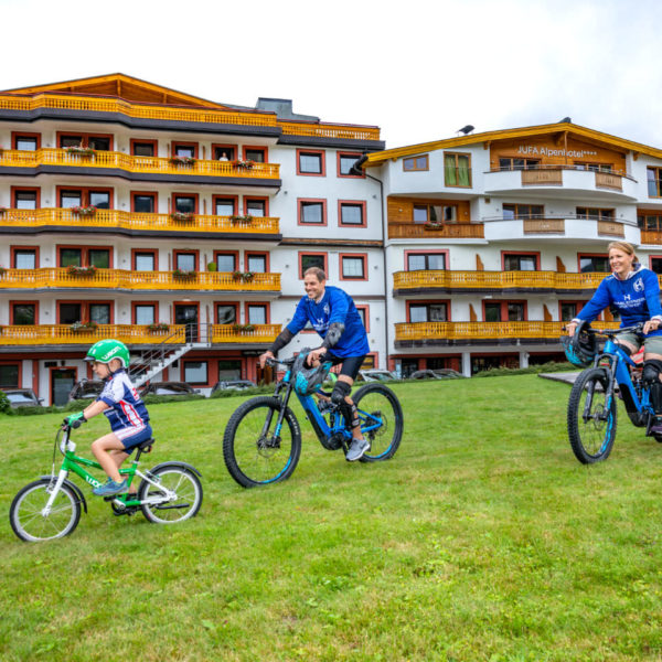 Sie sehen eine Familie beim Mountainbiken in Saalbach vor dem JUFA Alpenhotel Saalbach****. Der Ort für erholsamen Familienurlaub und einen unvergesslichen Winter- und Wanderurlaub.